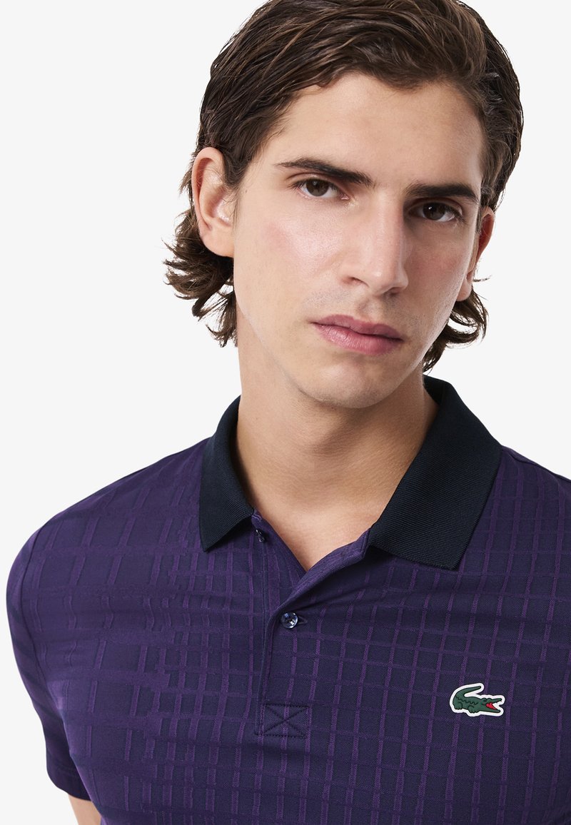 Polo shirt a maglia viola con una trama a reticolo, colletto nero e un piccolo logo del coccodrillo Lacoste sul petto sinistro.