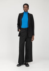 Zwarte op maat gemaakte blazer over een felblauwe coltrui, gecombineerd met zwarte wijde broek en stevige zwarte schoenen. Simpele, gladde textuur.