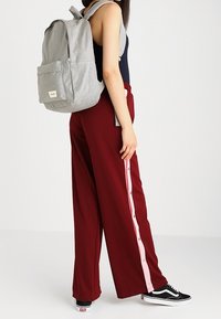 Sac à dos gris avec fermetures éclair et poche avant, porté sur un débardeur noir, associé à un pantalon large bordeaux avec des boutons roses sur le côté.
