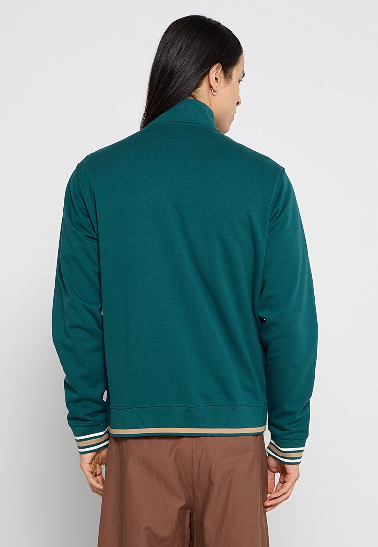 Felpa teal con colletto alto, polsini e orlo a coste, caratterizzata da strisce bianche e dorate. Tessuto liscio, vestibilità comoda e design della parte posteriore semplice.