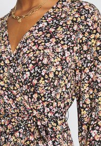 Czarna sukienka w kwiaty o fasonie kopertowym, z dekoltem w serek, z wyrazistymi kwiatami w kolorze żółtym, różowym i białym, z teksturowanej tkaniny oraz z wiązanym paskiem w talii.