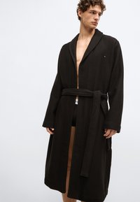 Melna vafeļu adījuma peldmētelis ar jostu viduklī, ar šāla apkakli un garām piedurknēm. Smalks logotipa detalizējums kreisajā krūtīs.