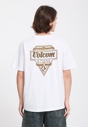 Jeune personne aux cheveux mi-longs portant un t-shirt blanc Volcom Stone avec un grand logo marron et un graphisme dans le dos.