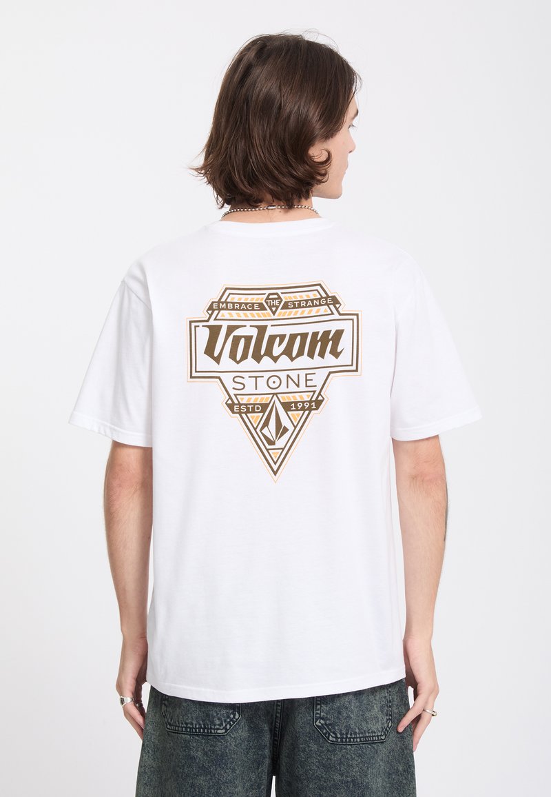 Jeune personne aux cheveux mi-longs portant un t-shirt blanc Volcom Stone avec un grand logo marron et un graphisme dans le dos.