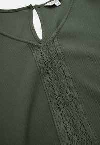 Dunkelgrüne Bluse mit einem Schlüssellochausschnitt, strukturiertem Stoff und einem Spitzenpaneel entlang der Mitte für zusätzlichen Detailreichtum.