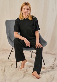 Frau mit blondem Haar, die ein schwarzes Outfit trägt, sitzt barfuß auf einem grauen modernen Stuhl vor einem beigen Stoffhintergrund.