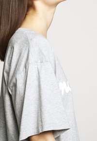 Profilvy av en person med långt hår som bär en ljusgrå oversized t-shirt med en vit logotyp på Ärmen, neutral bakgrund.