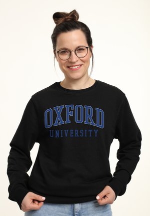 Smilende kvinde med briller og hår sat op i en knold iført sort Oxford University sweater og lyseblå jeans mod en ensfarvet baggrund.