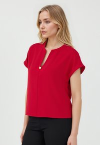 Haut rouge à manches courtes avec un col en V et des détails de boutons à l'avant. Fabriqué en tissu lisse, il a une coupe décontractée et des lignes épurées.