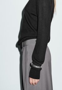 Maglione nero con maniche lunghe abbinato a pantaloni grigi. Un bracciale d'argento adorna il polso, enfatizzando un design elegante e minimalista.