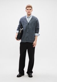 Gray fluffy cardigan with buttons, layered over a light blue striped shirt, paired with black trousers and black shoes; holding a black bag.