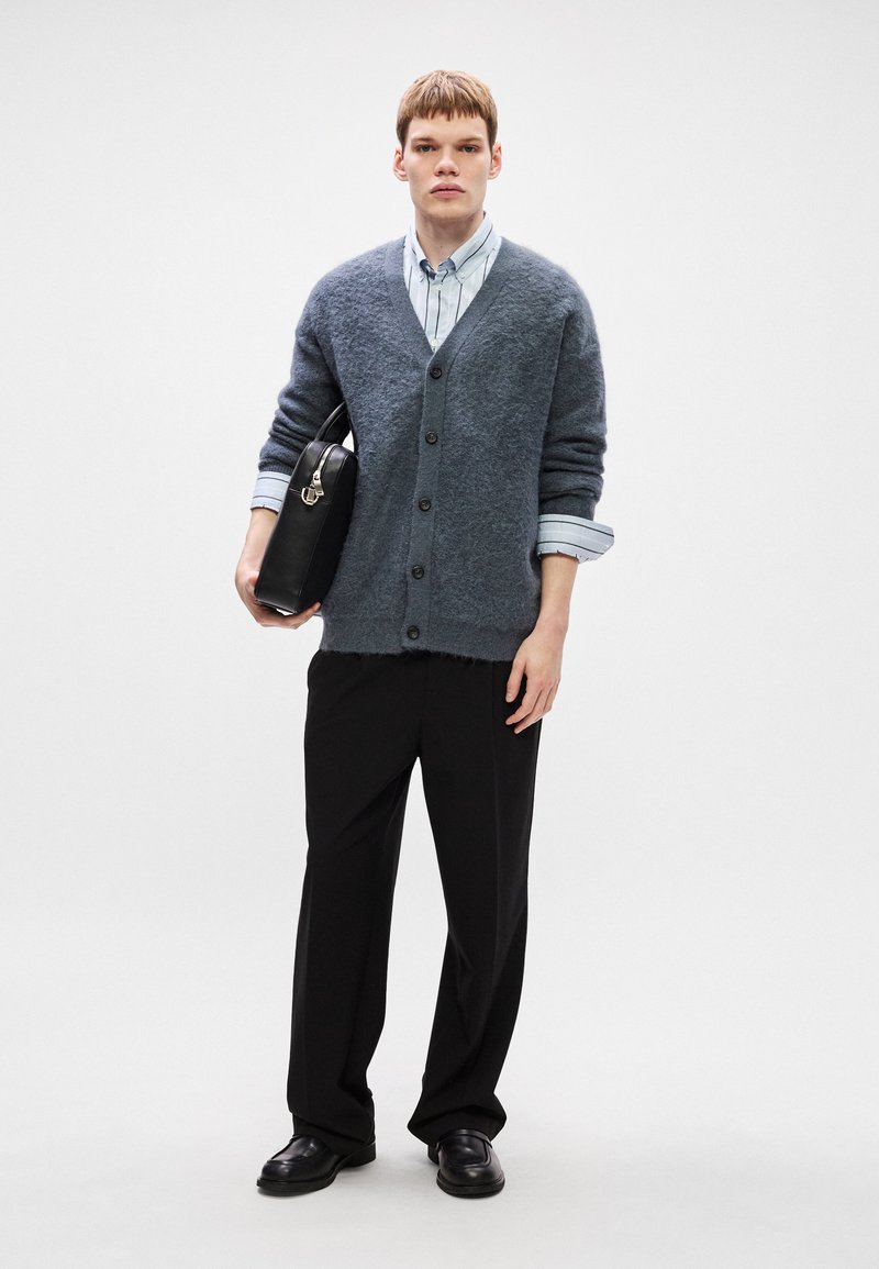 Gray fluffy cardigan with buttons, layered over a light blue striped shirt, paired with black trousers and black shoes; holding a black bag.