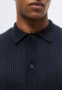 Κοντινό ενός ατόμου που φοράει σκούρο μπλε πλεκτό πόλο με κάθετες υφασμάτινες ρίγες και δύο μαύρα κουμπιά κλεισμένα στο γιακά.