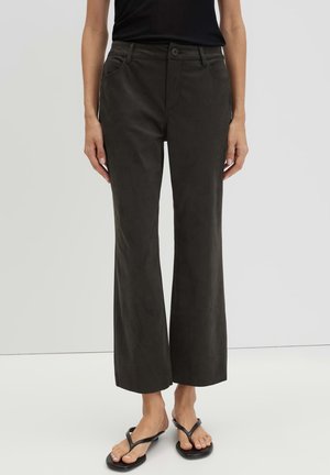 Persona che indossa pantaloni larghi marrone scuro tagliati sopra la caviglia e sandali infradito neri, in piedi davanti a uno sfondo chiaro e uniforme.