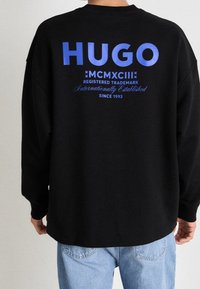 Svart sweatshirt med en stor blå "HUGO"-logotyp på ryggen, med ytterligare text. Tyget verkar mjukt och texturerat, med en avslappnad passform.