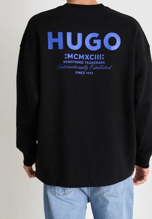 Sudadera negra con un gran logo azul de "HUGO" en la parte posterior, junto con texto adicional. La tela parece suave y con textura, con un corte relajado.