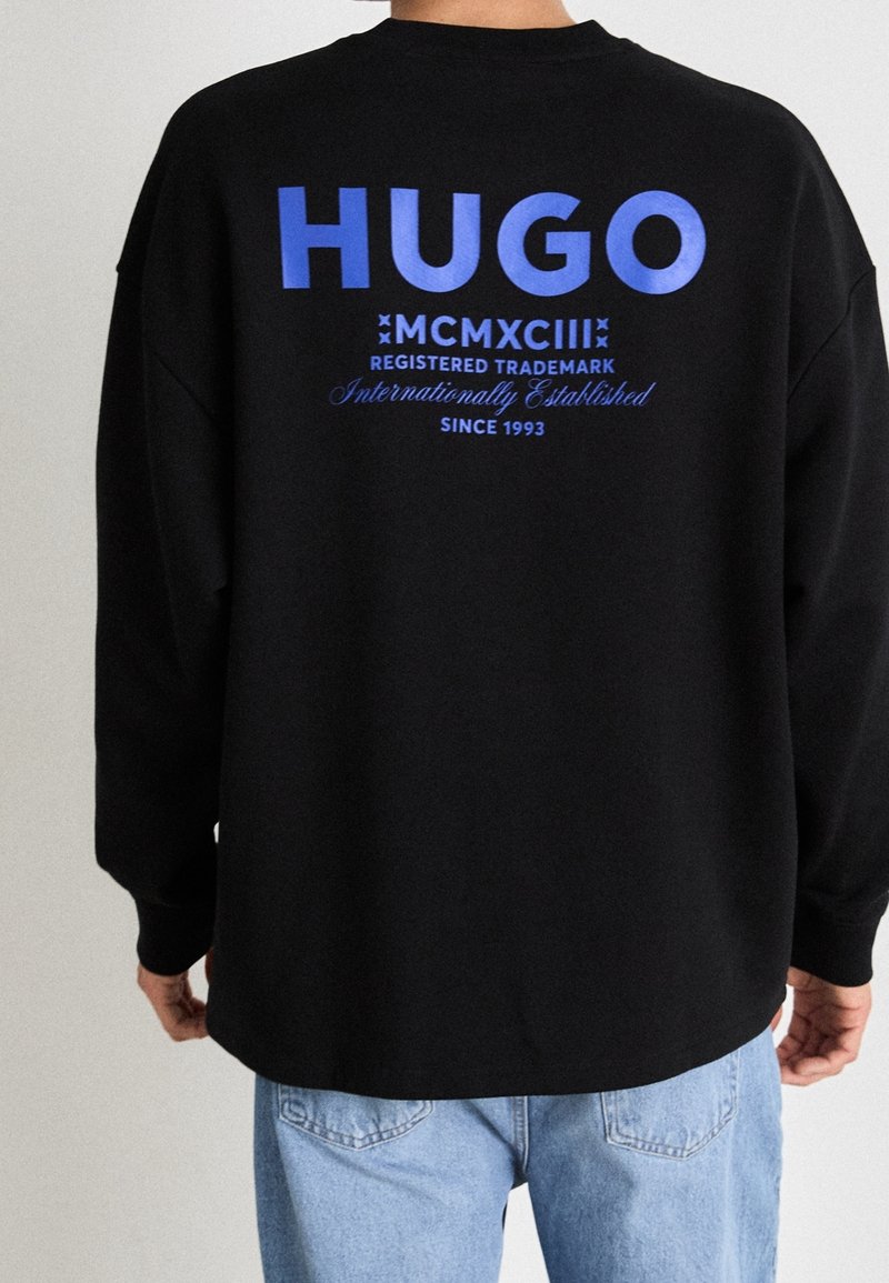 Svart sweatshirt med en stor blå "HUGO"-logotyp på ryggen, med ytterligare text. Tyget verkar mjukt och texturerat, med en avslappnad passform.