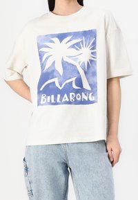 Off-white bomullst-shirt med en blå och vit grafik av palmer och sol med texten "BILLA BONG" längst ner. Enkel, avslappnad passform.