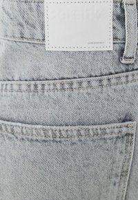 Harmaa farkkukangas, jossa on teksturoidu viimeistely, takatasku, valkoinen nahkalappu ja kontrastitikkaukset saumoissa.