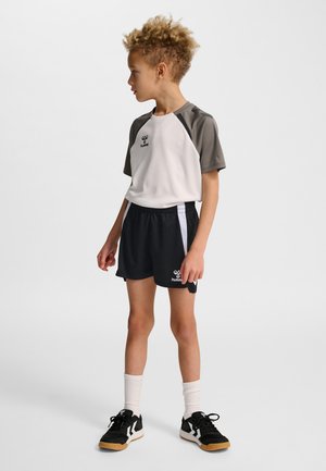 Kind mit lockigem Haar steht und schaut nach links, trägt ein weiß-graues Sportshirt, schwarze Shorts mit weißen Streifen, weiße Socken und schwarze Sneakers.