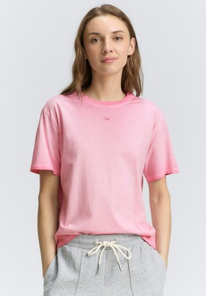 Camiseta de algodón rosa con mangas cortas, cuello redondo y un pequeño detalle de logo en el pecho. Combinada con pantalones de chándal grises con cordón ajustable.