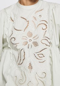 Weißes Blusenshirt mit aufwendiger floral gefertigter Lochstickerei auf der Brust und puffigen langen Ärmeln.