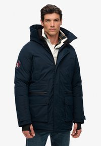 Veste isolée bleu marine avec capuche, fermeture éclair à l'avant et plusieurs poches. Présente une doublure à carreaux et un badge logo sur la manche.