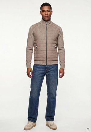 Mann trägt eine beige gesteppte Jacke mit Reißverschluss, blaue Jeans und beige Sneakers, steht frontal vor einfarbigem Hintergrund.