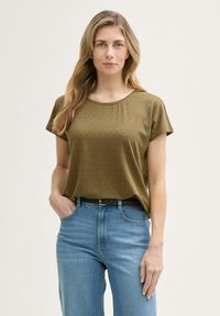Cămașă scurtă de culoare verde măslin, cu un model texturat, guler rotund, croială lejeră, purtată împreună cu blugi din denim de culoare bleu deschis și curea neagră.