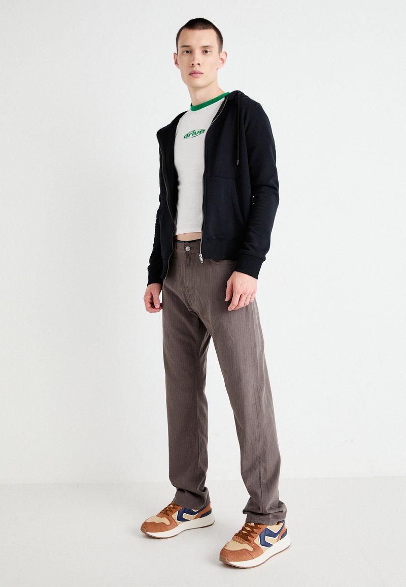 Sort zip-up hoodie, hvid T-shirt med grøn kant, brune stribede bukser og beige sneakers med blå detaljer.