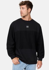Schwarzer Sweatshirt mit einem strukturierten Panel über der Brust, gerippten Bündchen und Saum. Mit einem kleinen Logopatch auf der Vorderseite.