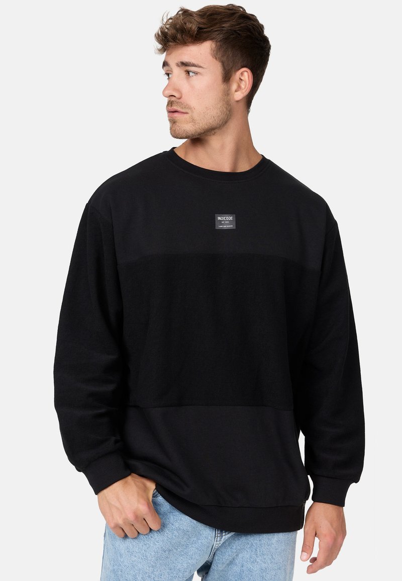 Schwarzer Sweatshirt mit einem strukturierten Panel über der Brust, gerippten Bündchen und Saum. Mit einem kleinen Logopatch auf der Vorderseite.