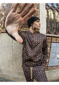 Chándal marrón con estampado y detalles de logo, chaqueta con cremallera, pantalones con cordón, hecho de tela suave; combinado con un gorro de lana negro.