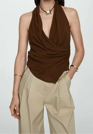 Kvinde iført en brun draperet haltertop, beige højtaljede plisserede bukser, gyldt firkantet vedhæng og gyldne armbånd på begge håndled.