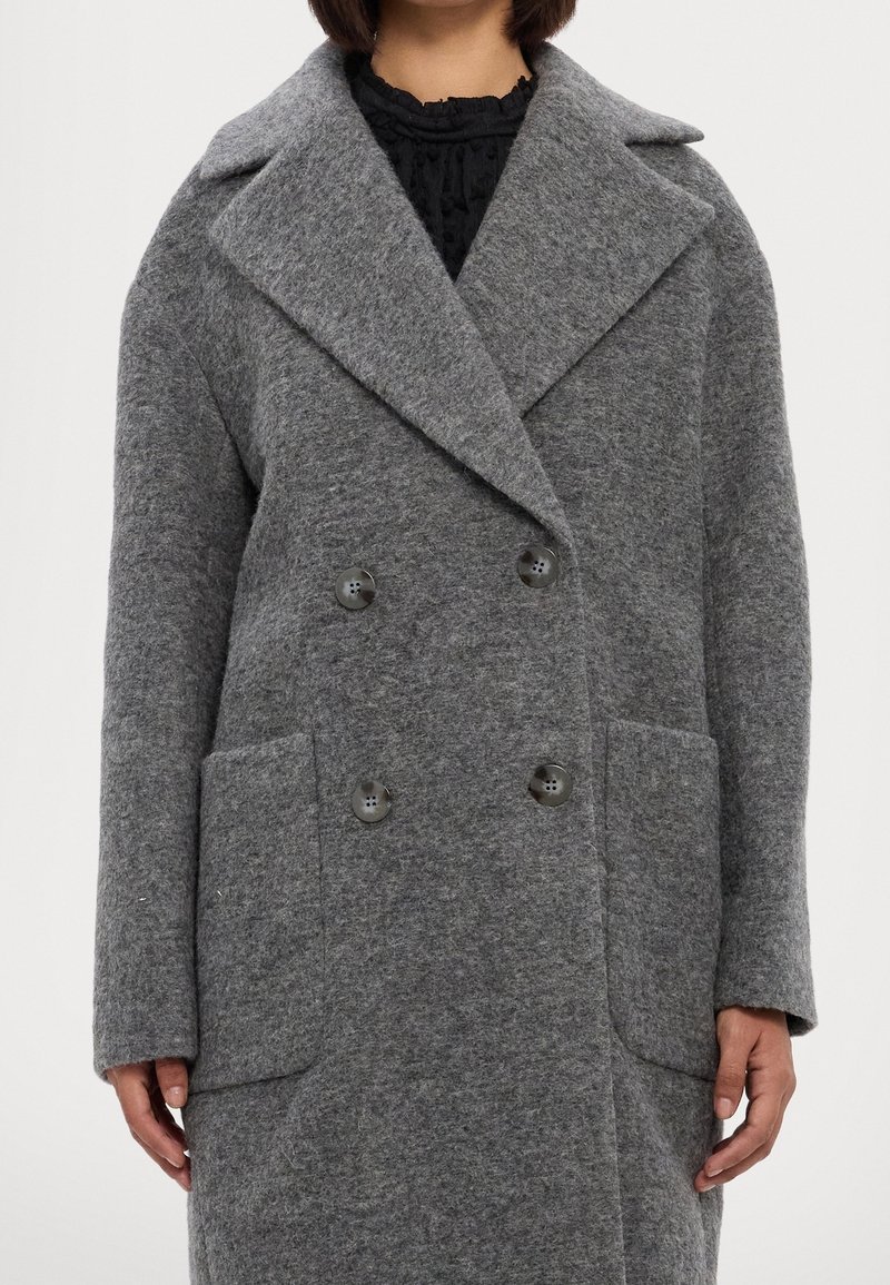 Personne portant un manteau en laine gris croisé avec de larges revers et de grandes poches avant sur un haut noir texturé.
