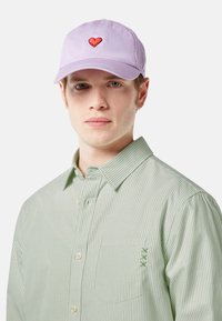 Lila baseballsapka piros szív hímzéssel az elején, egy zöld és fehér csíkos gombos inggel párosítva.