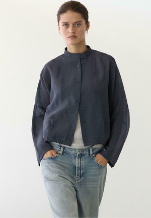 Femme aux cheveux bruns attachés en arrière, portant une veste sombre boutonnée sur un haut gris et un jean bleu clair, mains dans les poches, expression neutre.