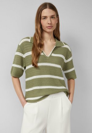 Vrouw met lang bruin haar draagt een groen-wit gestreepte gehaakte trui met korte mouwen en witte hoge taillebroek, handen in de zakken.