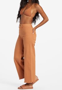 Pantalones de tiro alto y pierna ancha en un cálido tono terracota, con una tela texturada y un dobladillo sin rematar. Combinados con un sujetador de bikini marrón.