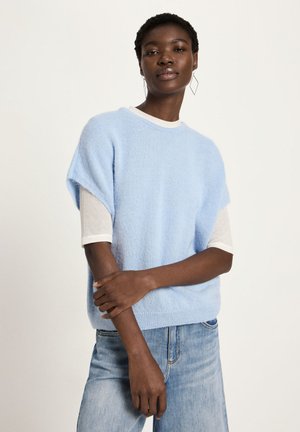 Woman wearing light blue short-sleeve sweater over white long-sleeve shirt and light blue jeans, standing with arms crossed against plain background.