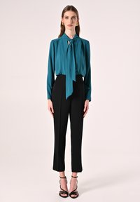 Blusa di seta verde acqua con un nodo al collo, abbinata a pantaloni neri a vita alta, completata da tacchi neri con cinturini.
