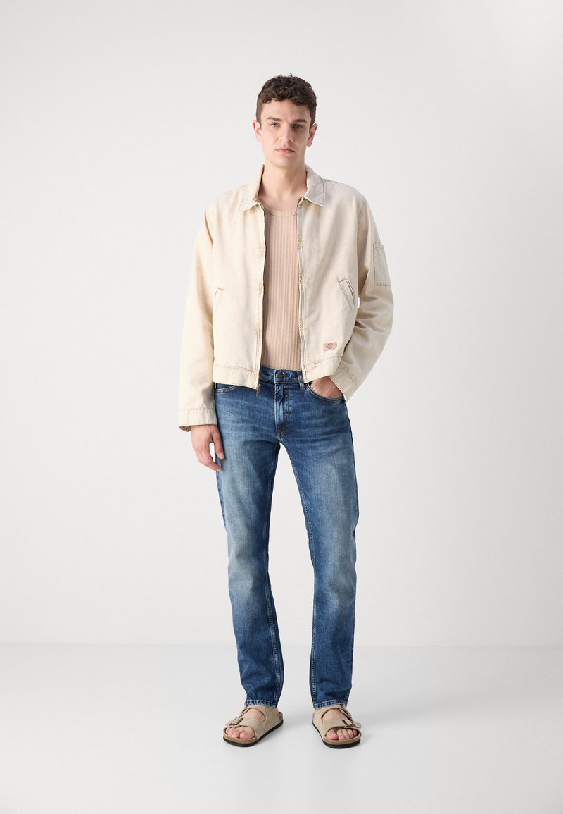 Beige Jeansjacke mit einem Reißverschluss vorne, hellbrauner Rippsweater, blaue gerade geschnittene Jeans und sandfarbene Sandalen auf einem einfarbigen Hintergrund.