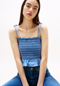 Lyseblå smocket topp i kort lengde med tynne stropper bundet på skuldrene, med en volangkant. Kombinert med høytlivede mørkeblå jeans.