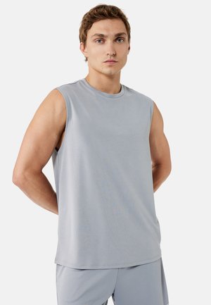 Młody mężczyzna z jasnobrązowymi włosami, ubrany w bezrękawnik sportowy w jasnoszarym kolorze i dopasowane szorty, stoi z rękami za plecami.