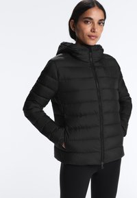 Nő áll, kezét a zsebében tartva, fekete kapucnis steppelt dzsekit és fekete nadrágot visel, egyszínű fehér háttér előtt.