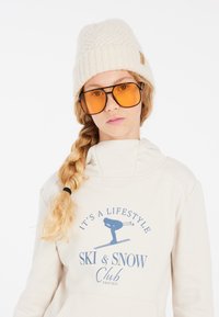 Felpa crema con grafica da sci e testo blu, completata da un berretto lavorato bianco e occhiali da sole grandi con lente gialla.