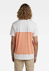 Hvid bomulds t-shirt med et orange og hvidt stribet design på ryggen. Korte ærmer og rund halsudskæring.