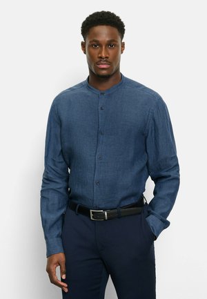 Mann trägt ein blaues Hemd mit Stehkragen und Knöpfen sowie dunkle Hose mit schwarzem Gürtel, steht vor einem schlichten weißen Hintergrund.