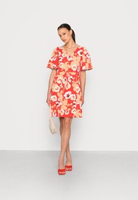 Vestido floral vermelho com mangas curtas e esvoaçantes, com cintura ajustada e decote em V. Combinado com saltos abertos vermelhos e uma mala bege.