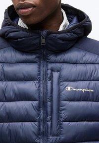 Κοντινό πλάνο ενός προσώπου που φοράει μπλε σκούρο καπιτονέ μπουφάν Champion με κουκούλα και λευκό λογότυπο στο στήθος, με το φερμουάρ τελείως κλειστό.